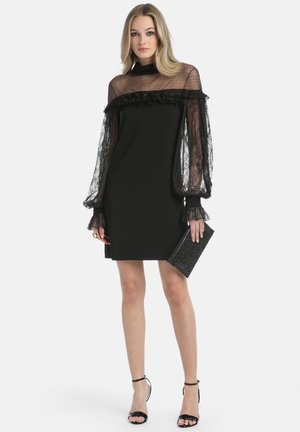 Zwart jurk met doorzichtige, stippenkant mouwen en ruches bij de halslijn, met een aansluitende silhouet en gladde stof. Houdt een zwarte clutch vast.