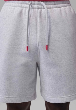 Hellgraue Shorts mit Kordelzug und weißen Bändern mit roten Enden, getragen von einer Person vor einem einfachen Hintergrund.
