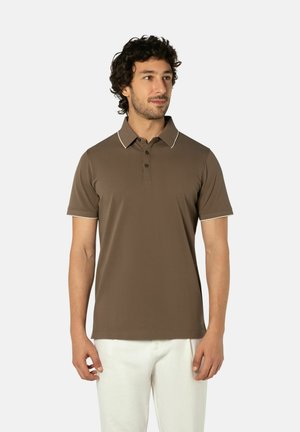 Man draagt een bruin poloshirt met korte mouwen en contrasterende biezen aan de kraag en een witte broek, staat en kijkt naar rechts.