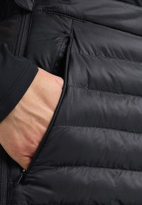 Zwarte geïsoleerde jas met horizontale stiksels, voorzien van een ritszak aan de zijkant. Het materiaal heeft een gladde, glanzende textuur.