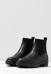 Botas de tobillo negras de cuero con lados de tela acanalada, suelas texturizadas resistentes y tiradores para ponerlas y quitarlas fácilmente. Diseño elegante y moderno para versatilidad.
