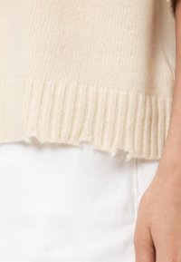 Maglione lavorato a maglia di colore crema; presenta un orlo a coste con bordi sfrangiati, realizzato in un materiale morbido e testurizzato. Una mano poggia su un tessuto bianco sottostante.