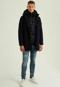 Jūrinės spalvos pūkinė striukė užsivilkta ant juodos užtrauktuku užsegamos striukės, derinama su dėvėtais šviesiai mėlynais džinsais ir baltais sportbačiais. Minkštos tekstūros, elegantiškas dizainas.