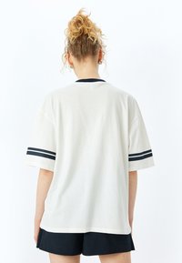 Hvid bomulds T-shirt med korte ærmer, der har en marineblå krave og to marineblå striber på hver ærme. Blød struktur, oversized pasform.