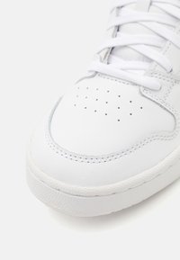 Valkoinen nahkakenkä, jossa on pyöreä kärki, rei'itetty yksityiskohta ja litteät valkoiset nauhat. Kumipohja, jossa on teksturoitu pinta. Saumasauma näkyvissä.