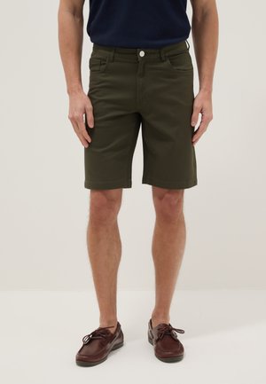 Hombre con pantalones cortos verde oscuro hasta la rodilla, camisa azul marino y zapatos de cuero marrón con cordones, de pie frente a un fondo liso.