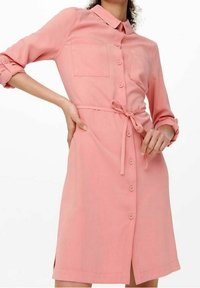 Abito rosa con bottoni, collo, due tasche sul petto e vita legata. Il tessuto sembra morbido con maniche arrotolate e spacchi laterali.