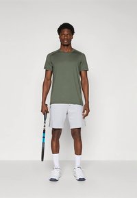 Zelené atletické tričko s krátkým rukávem, šedé šortky, bílé ponožky, drží černomodrý tenisový raketu. Jednoduchý design, lehká látka.