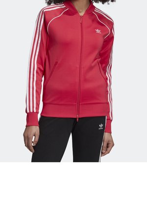 Kobieta w czerwonej kurtce sportowej Adidas z białymi paskami na rękawach i czarnych spodniach sportowych Adidas na jednolitym tle.