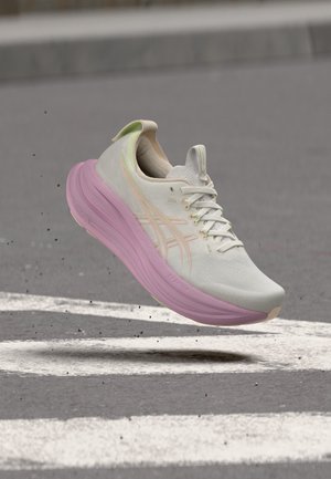 Chaussure de course blanche et rose pâle avec lacets, en l'air au-dessus d'une route asphaltée avec des lignes blanches peintes.