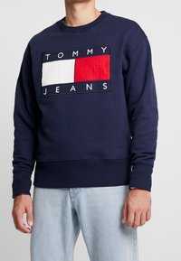 Hombre vistiendo un sudadera azul marino con un gran logo rectangular de Tommy Jeans en blanco y rojo, combinado con unos vaqueros de denim azul claro.