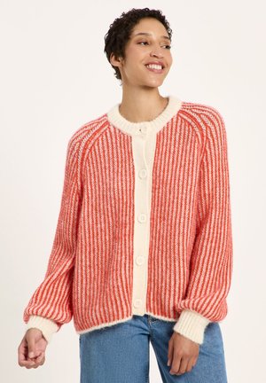 Frau, die lächelt, trägt eine rot-weiß gestreifte gestrickte Strickjacke mit Knöpfen, kombiniert mit blauen Jeans vor einem einfarbigen hellen Hintergrund.
