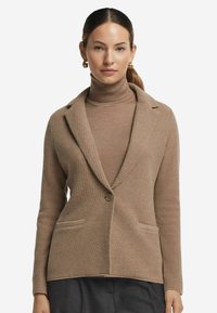 Beige stickad blazer med texturerad tyg, slagkrage, en knappstängning och två framfickor. Bärs över en matchande polotröja.