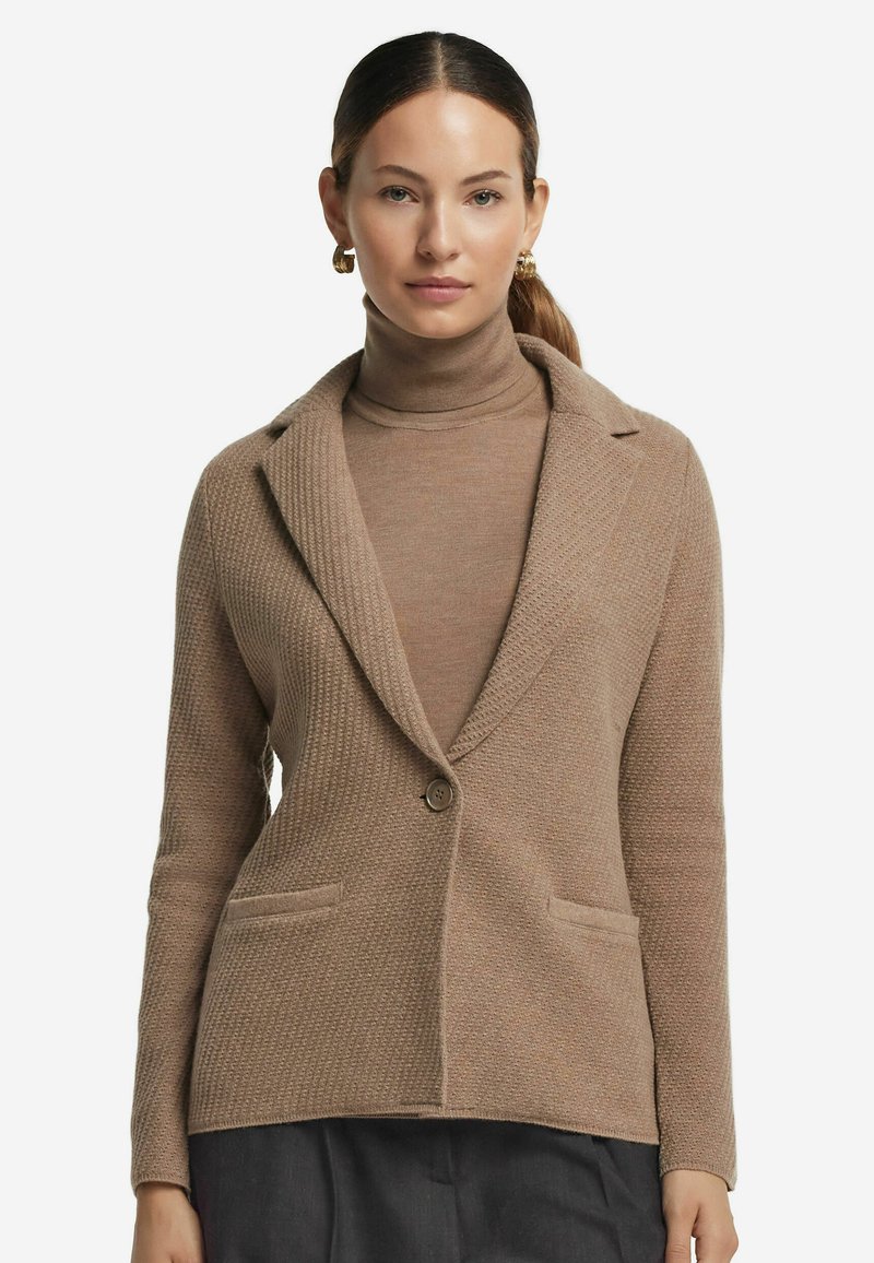 Beige stickad blazer med texturerad tyg, slagkrage, en knappstängning och två framfickor. Bärs över en matchande polotröja.