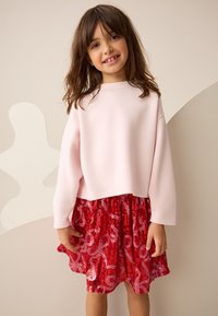 Felpa corta rosa chiaro con maniche lunghe abbinata a una gonna rossa con paillettes con motivo a vortice e orlo arricciato.