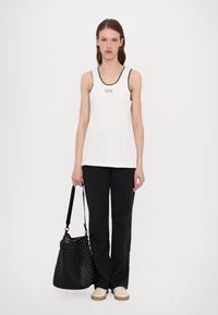 White sleeveless top with black trim and embroidered "MM" logo, paired with black trousers and a large woven black handbag.