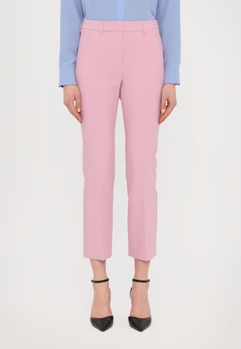 Femme portant un pantalon rose pastel sur mesure longueur cheville, associé à des escarpins noirs à bout pointu avec bride à la cheville et un chemisier bleu clair.