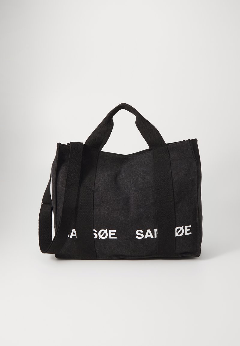 Saco de mão preto em lona com duas alças curtas e uma alça longa para ombro. Apresenta texto impresso em branco "SAMSØE" na parte da frente. Fecho de zipper no topo.