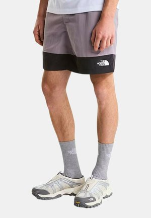 Hombre con pantalones cortos The North Face grises y negros, calcetines grises y zapatillas deportivas blancas y grises, de pie contra un fondo blanco.