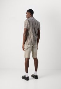 Camisa de manga curta com padrão de colmeia em tons de bege e castanho, combinada com calções bege claros e sapatos pretos.