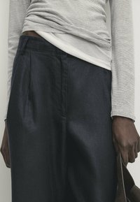 Chemise à rayures à manches longues en tissu doux, superposée sur un haut blanc, associée à un pantalon large à texture bleu foncé.