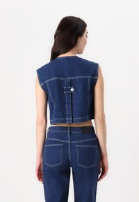 Gilet in denim blu scuro con fori ampi per le braccia, dettaglio delle maniche con bottone sul retro e cuciture a contrasto. Abbinato a pantaloni in denim blu scuro.