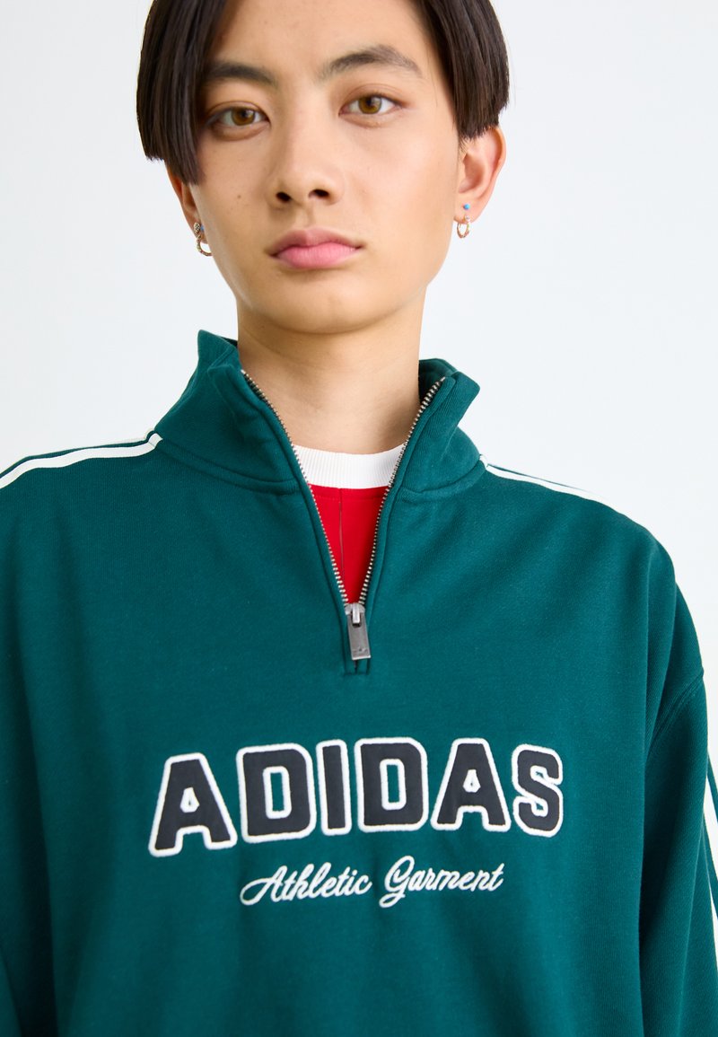 Sweat-shirt zippé vert avec le logo "ADIDAS", des rayures blanches sur les manches et un col montant. En dessous, une chemise rouge est visible.