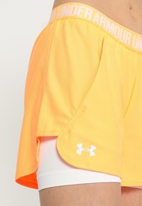 Shorts deportivos naranjas con una cinturilla ancha que presenta el texto 'UNDER ARMOUR' y un logo blanco en el dobladillo, superpuestos sobre unos shorts de compresión blancos.