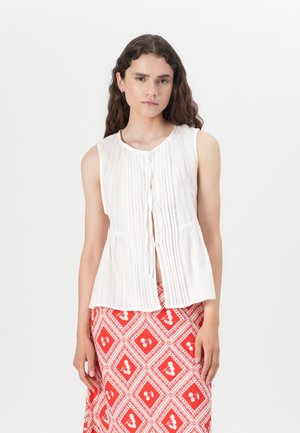 White, sleeveless top with vertical pleats, round neckline, and front button closure. Worn with a red patterned skirt.