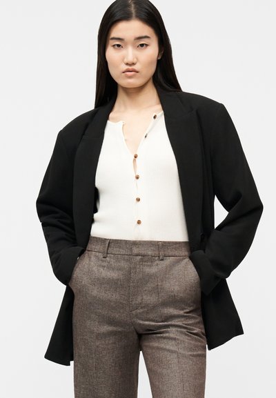 Femme aux longs cheveux noirs porte un blazer noir, un haut blanc boutonné et un pantalon marron sur mesure, debout les mains dans les poches.