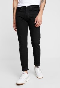 Jeans de mezclilla negra con un ajuste slim, diseño de cinco bolsillos y puños enrollados. Presenta cierre de botón metálico y cremallera. Llevados con zapatillas blancas.
