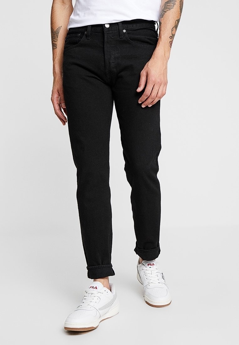 Jeans de mezclilla negra con un ajuste slim, diseño de cinco bolsillos y puños enrollados. Presenta cierre de botón metálico y cremallera. Llevados con zapatillas blancas.