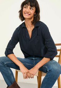 Marineblaues Langarmshirt mit V-Ausschnitt und einer Brusttasche, kombiniert mit blauen Jeans. Glattes Material, hochgekrempelte Ärmel und dezente Textur.