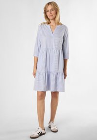 Blauw-wit gestreepte jurk met een V-hals, driekwart mouwen en een gelaagde volantzoom. Draagt witte sneakers.