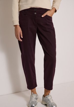 Personne portant un pantalon violet foncé et des baskets argentées, une main dans la poche et une chemise beige à manches longues visible à la taille.
