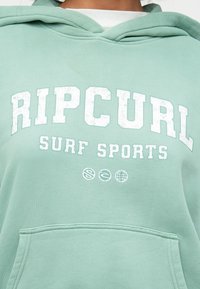 Mintgroene hoodie van zacht materiaal met een voorkantzak en witte vervaagde tekst "RIP CURL" en "SURF SPORTS."