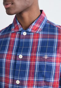 Camisa de cuadros roja y azul con cuello de botón, con botones blancos y un bolsillo en el pecho con un logo, fabricada con un tejido suave.