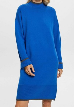 Vrouw draagt een lange blauwe gebreide trui-jurk met donkergrijze bies aan de manchetten, halslijn en zoom, staand voor een effen achtergrond.
