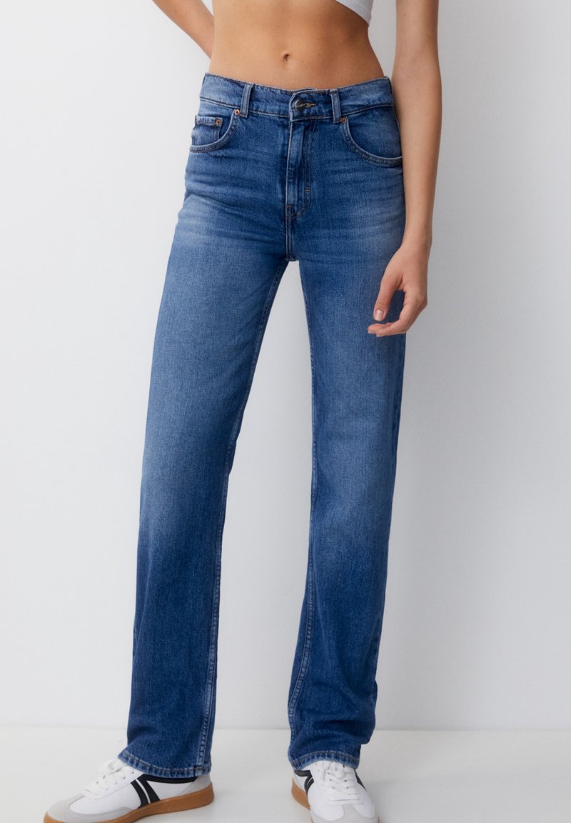 Jean en denim bleu à taille haute avec une coupe droite, cinq poches et un léger effet délavé sur les cuisses, accompagné de baskets blanches.