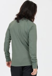 Person mit schulterlangen dunklen Haaren, die ein olivgrünes Langarmshirt und schwarze Hosen trägt, mit dem Rücken zur Kamera.