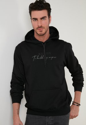 Crna hoodie s kapuljačom na vezanje, prednjim džepom i tiskanim tekstom. Izrađena od mekog materijala s rebrastim rukavima i rubom.