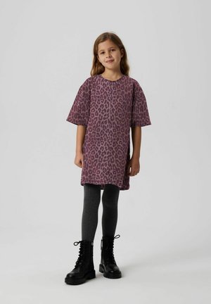 Mädchen steht in einem lila Leopardenmusterkleid, dunklen grauen Strumpfhosen und schwarzen Schnürkampfschuhen vor einem schlichten hellen Hintergrund.