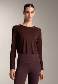 Nainen, jolla on taakse vedetyt hiukset, pukeutuneena hyvin istuvaan pitkähihaiseen croppitoppiin ja korkeavyötäröisiin leggings-housuihin tummanruskeana, seisoo vaaleanbeigeä taustaa vasten.