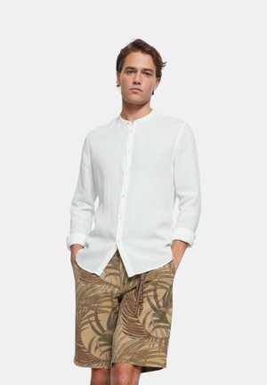 Giovane con le mani in tasca che indossa una camicia bianca a maniche lunghe con bottoni e pantaloncini beige a stampa tropicale fino al ginocchio.