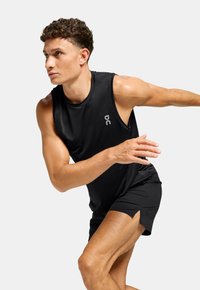 Canotta atletica nera con scollo rotondo e logo. Modello in movimento, indossa pantaloncini neri abbinati con spacchi laterali per maggiore flessibilità.