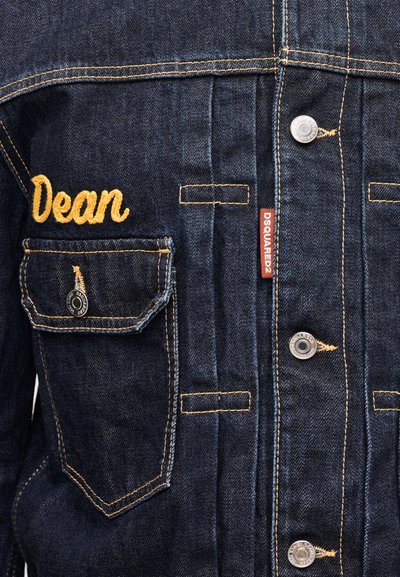 Chaqueta de mezclilla azul oscuro con costuras amarillas, nombre bordado "Dean" sobre el bolsillo izquierdo del pecho y etiqueta roja DSQUARED2 cerca de los botones.