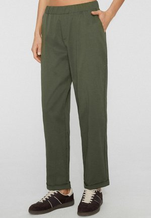 Person wearing olive green elastic waist pants with side pockets and black sneakers with white laces standing against a plain background.