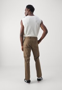 Brown chinos with a straight-leg fit, cotton blend material, and back pockets. Paired with a white sleeveless top and black sneakers.