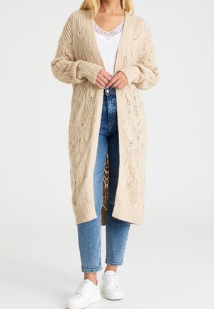 Femme portant un long cardigan beige en maille, un haut blanc bordé de dentelle, un jean bleu et des baskets blanches, debout devant un fond uni.