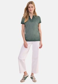 Polo shirt verde scuro a maniche corte con scollo a V, abbinato a pantaloni bianchi a gamba larga e sneakers chiare con dettagli verdi.